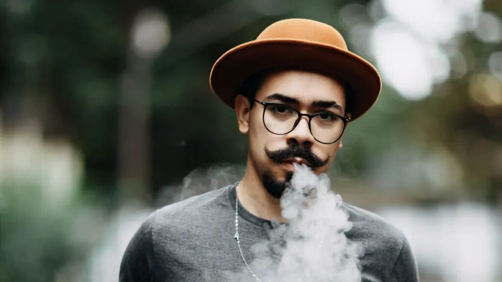 一个有胡子和山羊胡子的男人，穿着灰色的T恤，眼镜和软呢帽，呼出了一大羽毛。背景是一个模糊的户外环境，带有绿色植物。
